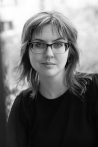 A black-and-white photo of a white person in their mid-30s with a shag haircut and glasses against an out-of-focus outdoor background. They are wearing a black shirt and smiling slightly.