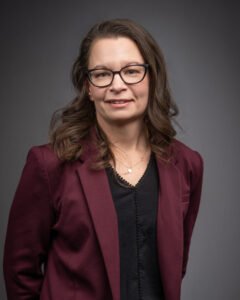 The author, a caucasian female in her early 40s with brown, curled hair just past her shoulders. She wears black framed cat-eyed glasses, a burgundy blazer, and a black top and stands in front of a gray backdrop.
