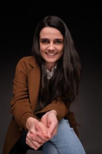 A white woman with long brown hair, leaning forward and smiling, in front of a black background. She’s wearing a white shirt and a brown coat.