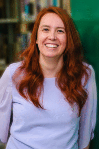 A photo of Brianna, a white woman with red wavy hair. She is standing in a library and smiling directly at the camera.