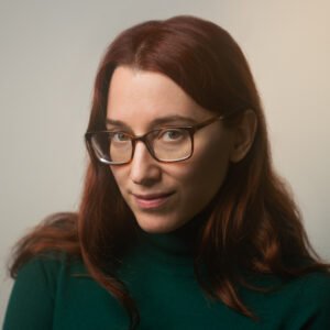 Photo shows a white woman with red-brown hair, green eyes, glasses with a brown frame, and a green turtleneck. She is shown from the chest up against a white background.