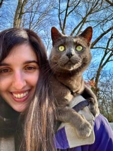 Photo of a white woman with dark hair and brown eyes, smiling while outside with trees in the background and a gray cat on her shoulder.