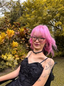A photo of the author, an Asian person with shoulder-length pink windswept hair and tattoos coming up their left arm, smiling into the camera in front of some foliage, wearing a black choker with a tiny silver heart in the middle and a detailed black dress.