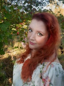 Photo of the author, a white person with long red wavy hair, standing in front of a tree with red berries during a summer day, wearing a white floral dress.