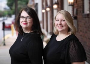 A picture of two white women in their mid-thirties, standing outside next to local businesses. The one on the left has shoulder-length brown hair, glasses, and blue eyes. She is shown from the chest up, smiling, and is wearing a black long-sleeve shirt. The one on the right has just above the shoulder blonde hair and blue eyes. She is shown from the chest up, smiling, and is wearing a short-sleeve black shirt.