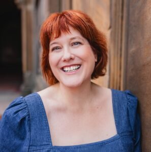 An outdoor photo of a white woman with blue eyes and red hair smiling in a denim dress.