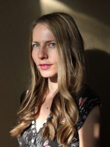 A photo of a white woman with long, light-brown hair, light green eyes, and a small smile, standing in front of a shadowed wall. She is wearing a black v-neck t-shirt with butterflies on it and a small silver skull necklace.