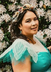 A photo of a person wearing a flower crown with dark brown hair and blue earrings and a blue dress with white petal-shaped sleeves and white lace front panel; in the background greenery and shrubbery with white and yellow flowers.