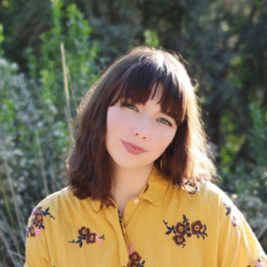 A light-skinned young woman with short shoulder-length brown hair and bangs, wearing a yellow collared shirt with embroidered flowers in front of a blurred green field.