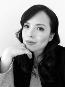 A black and white photo of a woman with long dark hair, she has her chin propped on her hand and is smiling.