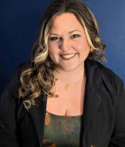 A photo of a smiling white woman with a blue background. She has long curled brown hair, and is wearing a black blazer.