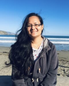 A photo of a smiling woman with long dark hair. She’s wearing glasses and a blue sweater and is sitting on the beach, with the sea in the background.