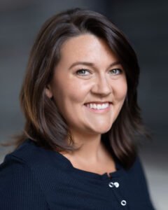 A picture of a white woman with shoulder-length brown hair and hazel eyes. She is shown from the chest up, smiling, and is wearing a black shirt.