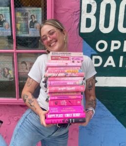 A picture of a smiling white woman with blonde hair and glasses, posing with a stack of books, in front of a bookstore window.