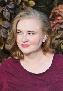 A white woman with blonde hair, wearing a maroon dress and silver ear cuff, looking slightly off camera while standing in front of some autumnal foliage.