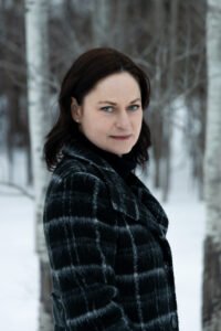 A woman with dark hair and blue eyes wearing a black and white plaid coat standing outdoors in a snow-covered forest.