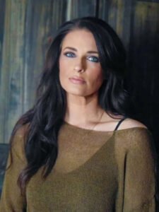 A.T. Rainach, a white woman with long, dark hair looks directly at the camera. She wears a bronze-brown top against a softly textured, muted background.