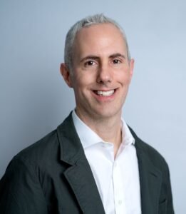 Male in gray blazer with white collared shirt smiling. Gray hair.