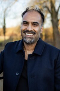 Ainkaran Sivaaji, a man with black hair and brown skin stands outdoors in an autumn countryside estate, in a dark blue long coat. He is looking at the viewer and has a bright smile.