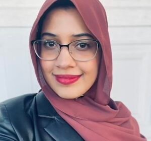 Hijabi woman with glasses, wearing a maroon hijab and maroon lipstick.
