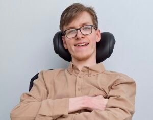 Alan Carrigan, a young brown-haired white man in a wheelchair with his arms crossed, wearing glasses and a beige shirt, smiling at the camera in front of a white wall.