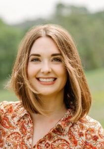 Aleese Lin, a white woman with a long bob hairstyle, stands in a field on a spring day, smiling brightly.