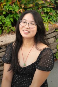 Alice Li, an Asian-American woman sits outside and smiles at the camera. She has glasses, long black hair, and is wearing a black dress with white polka dots. She is wearing a jade pendant hanging from a red string around her neck.