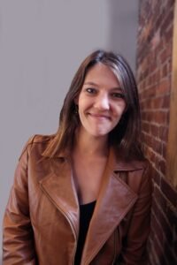 Alice Martin, a white woman with long brown hair sits in front of a brick wall. She is wearing a back top and a brown jacket. She is looking at the viewer with a slight smile.