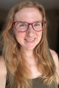 A close-up portrait photo of a smiling white woman with long red hair past the photo's edge, red glasses, and a green tank top.