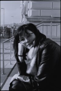 A black and white portrait of the author, a white woman with dark hair and bangs, wearing a leather jacket and sitting outside in Vienna. She has her chin propped on her fist and is smiling.