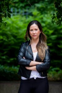 Author Amanda Connolly wears a leather jacket standing in front of a green row of trees.