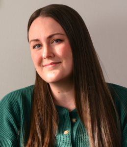Amanda Gayle, a white woman with medium brown hair and blue eyes, wearing a green, waffle-knitted shirt and making eye contact with the camera with a small, closed-lipped smile.
