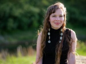 Amber Blaeser-Wardzala, a mixed Indigenous woman with long wavy hair, sits outdoors. She wears a sleeveless black mock neck and long earrings made from deer antlers.