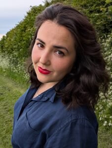 Amy Archer, a white woman with shoulder-length dark brown hair, stands outdoors wearing a navy blue shirt. She is looking over her shoulder at the camera.