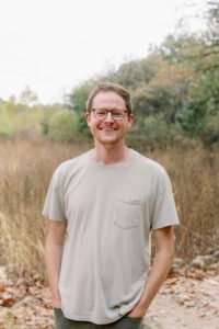 Andrew Forrester, a white man with glasses and light brown hair, standings outside against a backdrop of trees with his hands in his pockets.