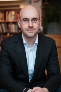 Andrew Reid is a white man with a bald head and glasses. He is wearing a blue shirt open at the collar and a dark suit jacket. He is sitting in a library and smiling at the camera.