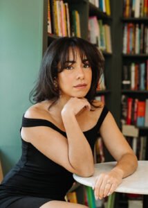 Andy C. Naranjo, a Latine woman with short black hair, sits in front of a blurred bookshelf with her elbow on a table and chin propped on a hand. Photo by Riley Glenn Photography.