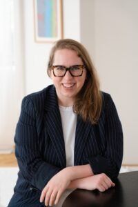 Anna McCallie, a white woman with dark blonde hair, sits in a brightly lit room with white walls. She is wearing a navy blue pinstriped blazer and has her arms crossed over one another as they rest on a table. She wears large tortoiseshell glasses and is smiling at the viewer.