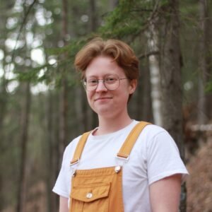 Photo of Ari Koontz, a white nonbinary person with short reddish hair, wearing a white t-shirt and mustard-yellow overalls along with round glasses. In the background is a blurred green and brown forest.