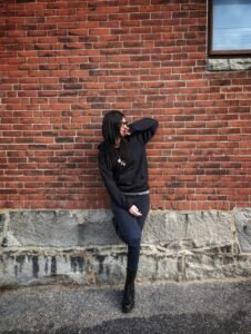 Person with dark hair wearing a black hoodie and black skinny jeans with boots leaning against a brick wall. They are looking away from the camera.