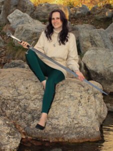 A white woman with long, dark hair (that's actually purple but looks brown in the photo) sits on a boulder in the middle of a creek. She's holding a large sword point down across her body and smiling. She's wearing green leggings and a cream cable knit sweater