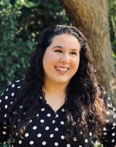 Ashley Tropea, an Egyptian-Italian woman with long, curly brown hair, stands in front of a tree. She is visible from the chest up and looks off into the distance.