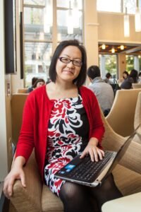 Betty Chang is an Asian woman with medium-length dark hair and glasses. She is sitting inside a cafe with a laptop on her lap. She is looking at the viewer and has a slight smile.