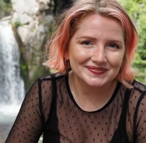 Betty Hawk, a white woman with pink highlighted hair, sits outside in front of a waterfall. She is looking at the viewer with a closed mouth smile.