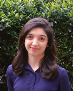 Bita Behzadi, a Middle Eastern woman with black hair stands in front of a green leafy background in a navy blue collared shirt smiling slightly at the viewer.