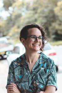 A photograph of Bori Cser, a white woman with curly brown hair cut in an asymmetrical pixie cut, round tortoiseshell glasses, a nose ring and three visible ear piercings. She stands outdoors with arms folded, wearing a patterned collared short-sleeved shirt in dark blue, light blue, orange and white zigzags and angular shapes. She is looking to the right and smiling. In the background are out of focus parked cars and trees.