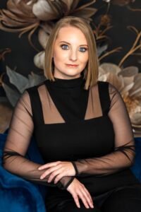 C.N. Vair, a white woman with a blonde bob, is seated in a blue velvet chair against a dark botanical backdrop.
