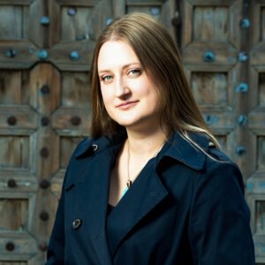 A woman with dark blonde hair smiles at the camera in three-quarter view. She is wearing a blue coat.