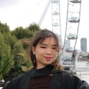 Camille Chong, a young looking Asian woman with long brown hair, standing before a grey sky. The photo is cropped to show her head to her shoulders. She is wearing a black gown that covers her shoulders, and she is smiling slightly.