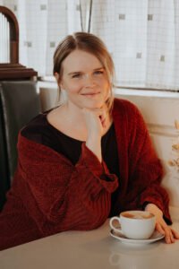 A white woman in her early 30s with dark blonde hair and blue eyes. She is sitting at a booth with a cup of coffee, wearing a dark purple top and a brown cardigan.